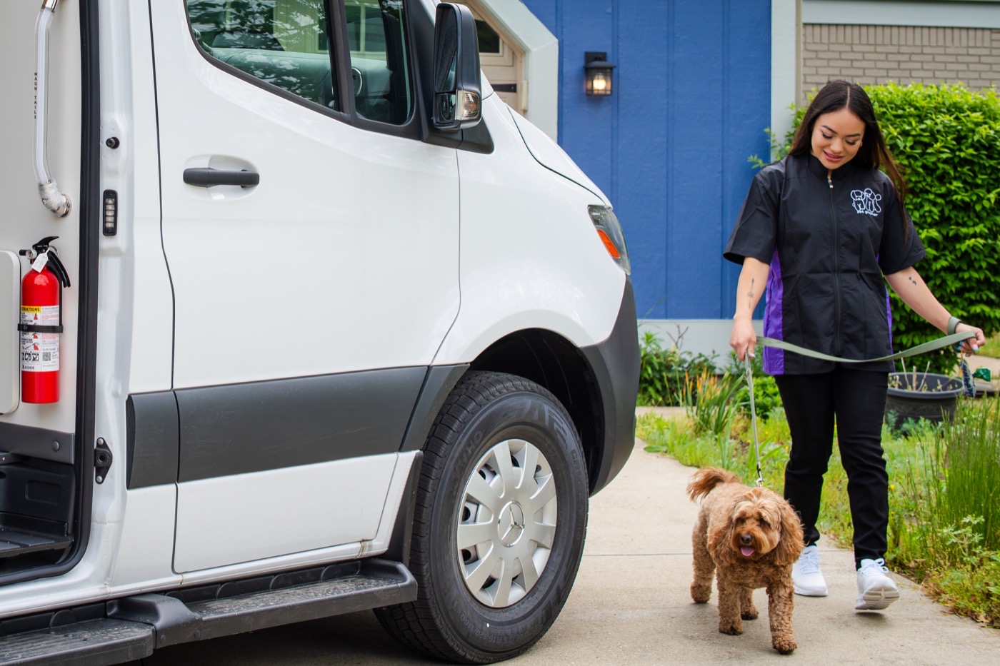 Mobile groomer walking a doodle toward a Wag'n Tails van on a residential driveway