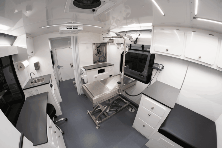 Interior of a white mobile medical laboratory with stainless steel workbench, cabinets, and overhead equipment mounted to the ceiling.