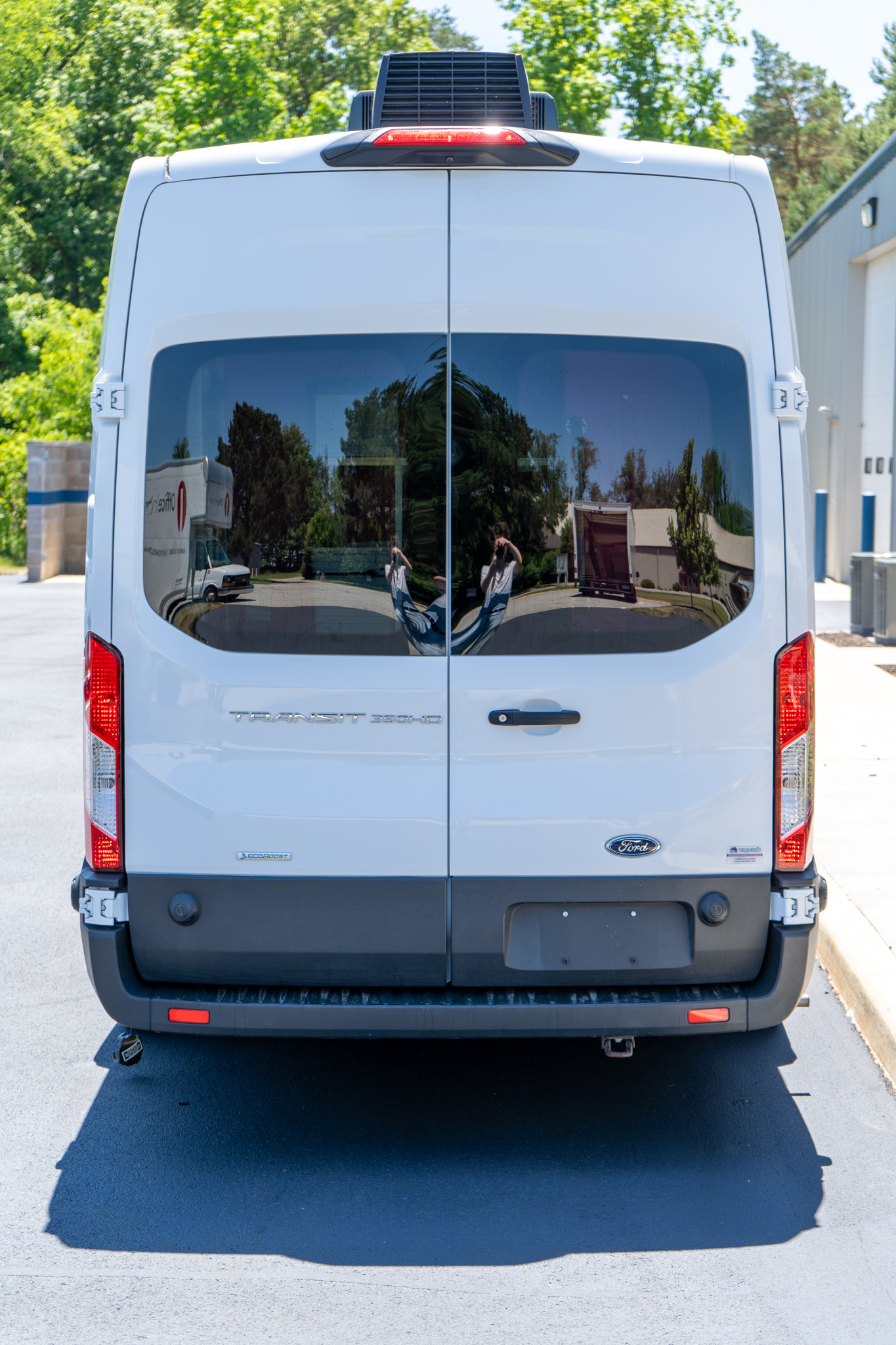 Rear view of a white Ford Transit van with dual back doors and tinted rear windows, reflecting trees and surroundings in the glass.