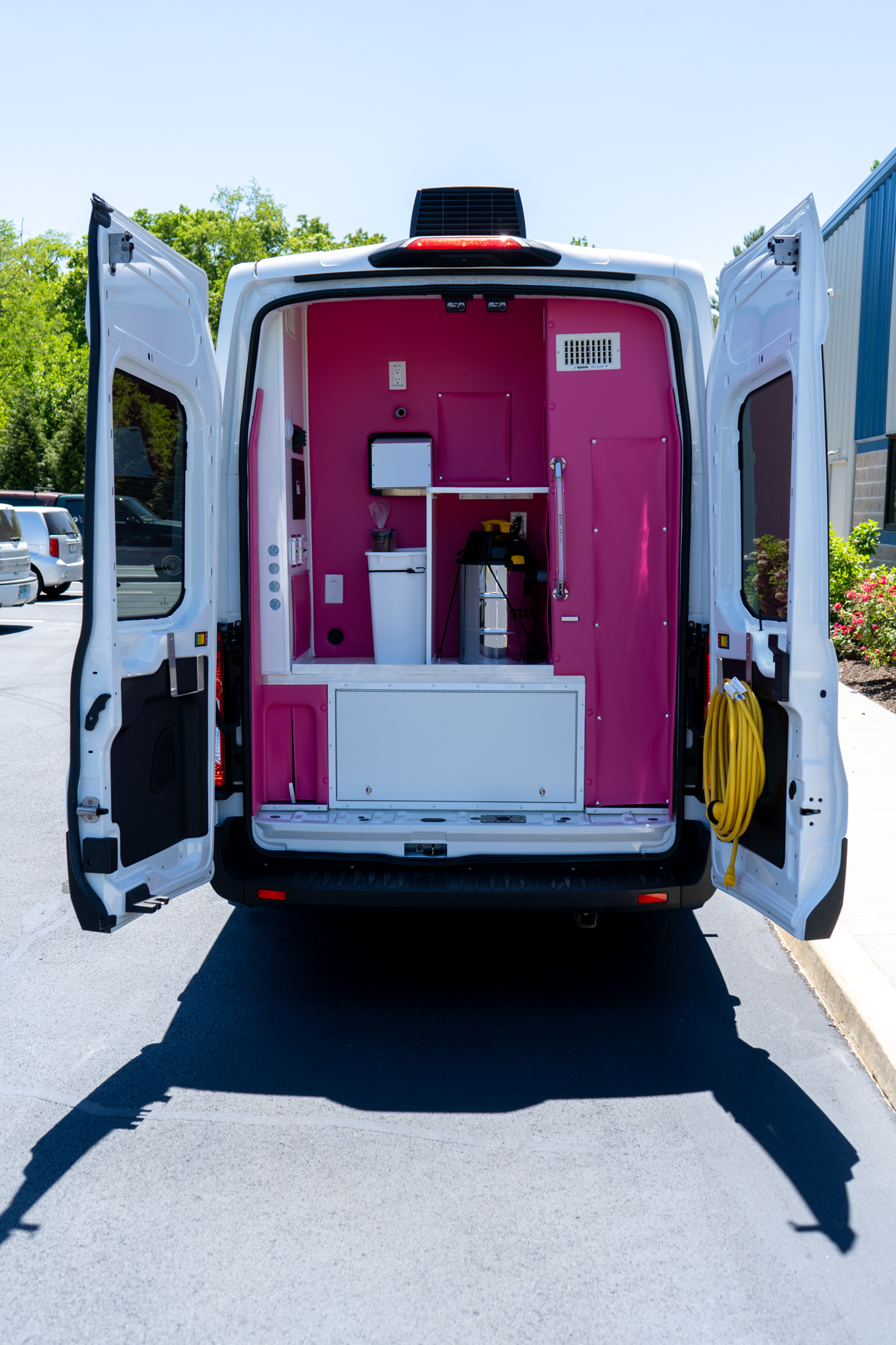 Open rear of a white van showing a bright pink interior workspace with shelves, a toolbox, and a coiled yellow hose nearby.