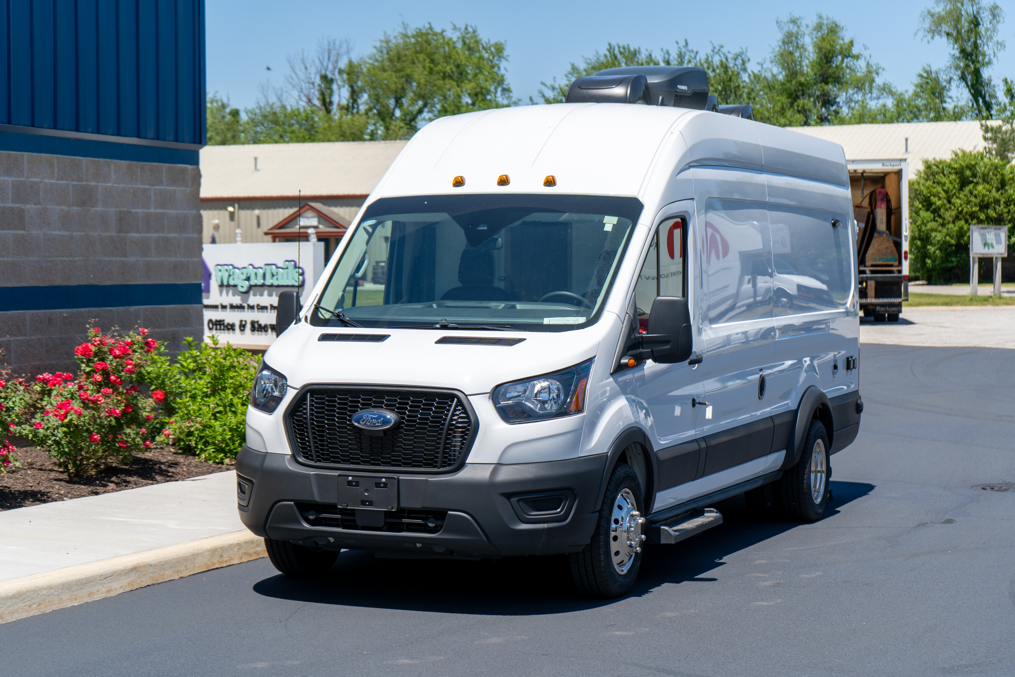 White cargo van parked near a blue building with a delivery truck in the background on a sunny day in a commercial area.