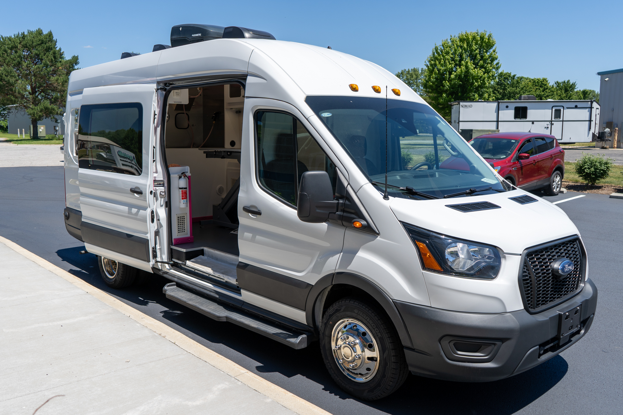 White camper van with its side door open, showing interior cabinets in a parking lot under a clear blue sky.