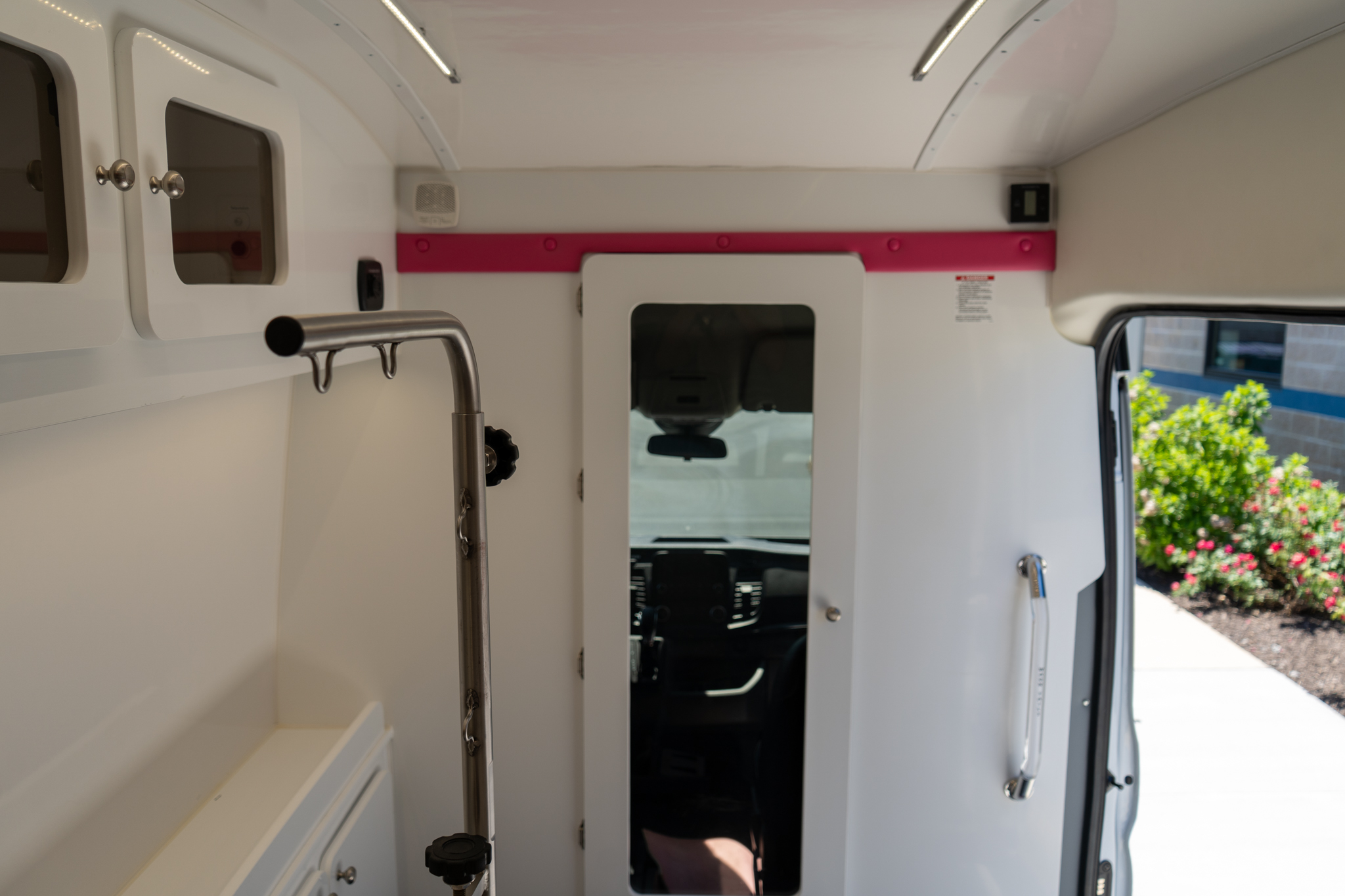 Interior of a white utility van with open cabinets and a pink horizontal strip along the wall, view toward the driver's area through a doorway.