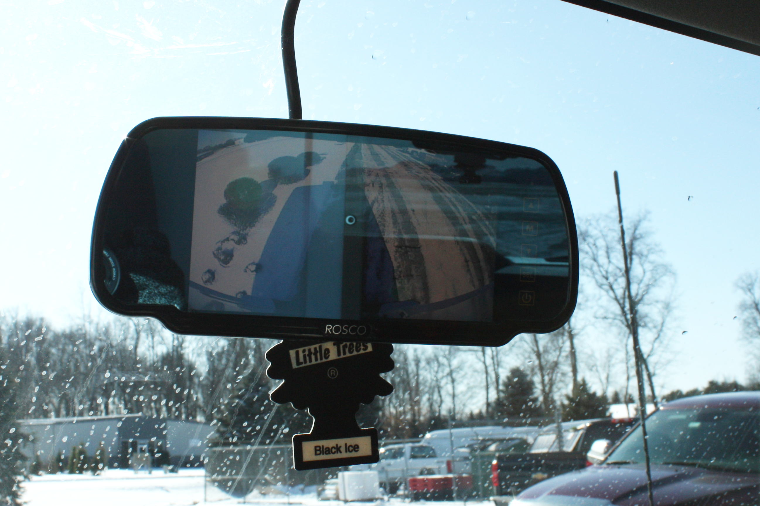 Rearview mirror with a Black Ice Little Trees air freshener hanging inside a car, snow outside the windshield.