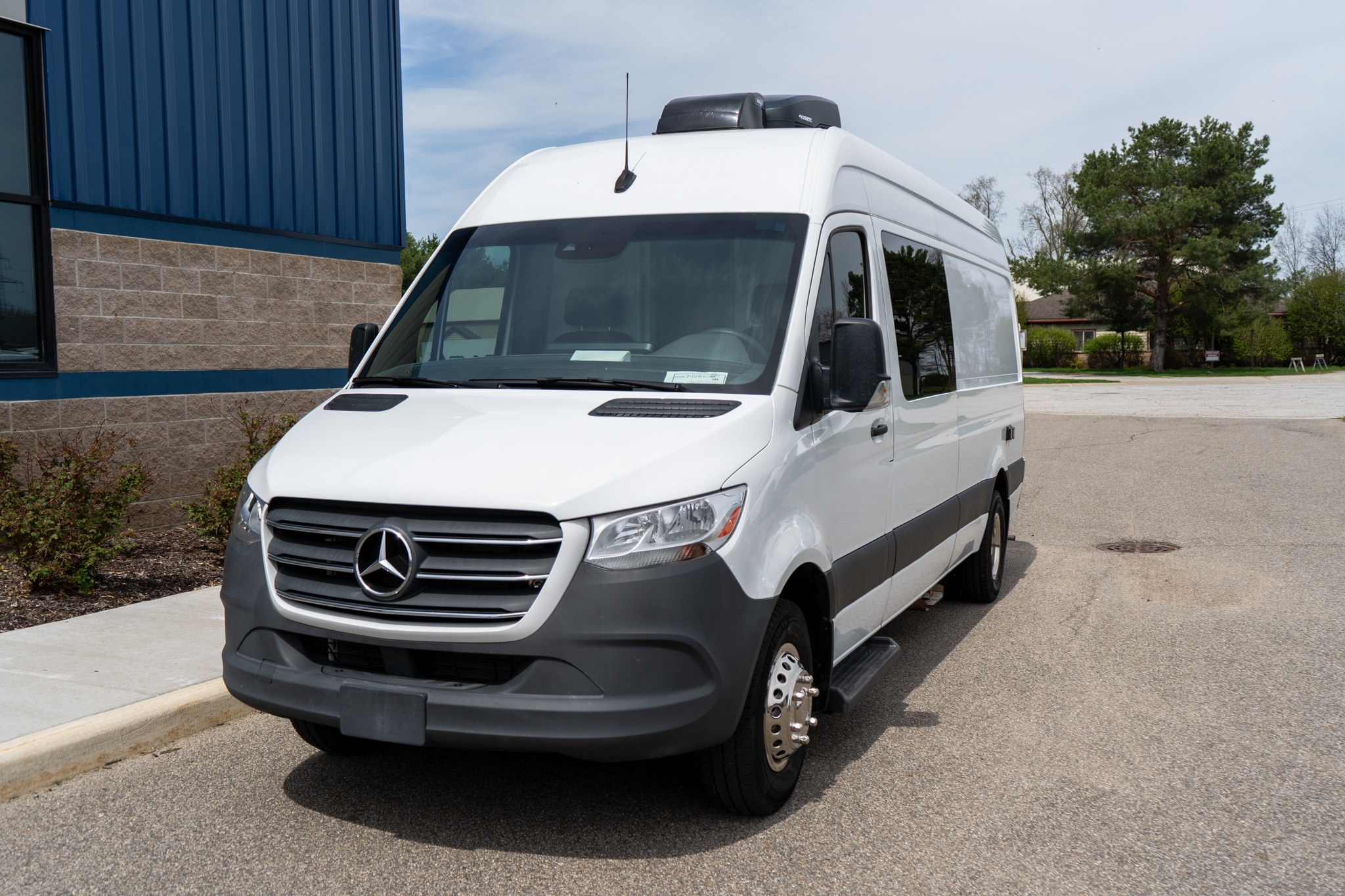 Front-left view of a white Mercedes-Benz cargo van parked in a lot beside a blue building, with a roof cargo box visible.