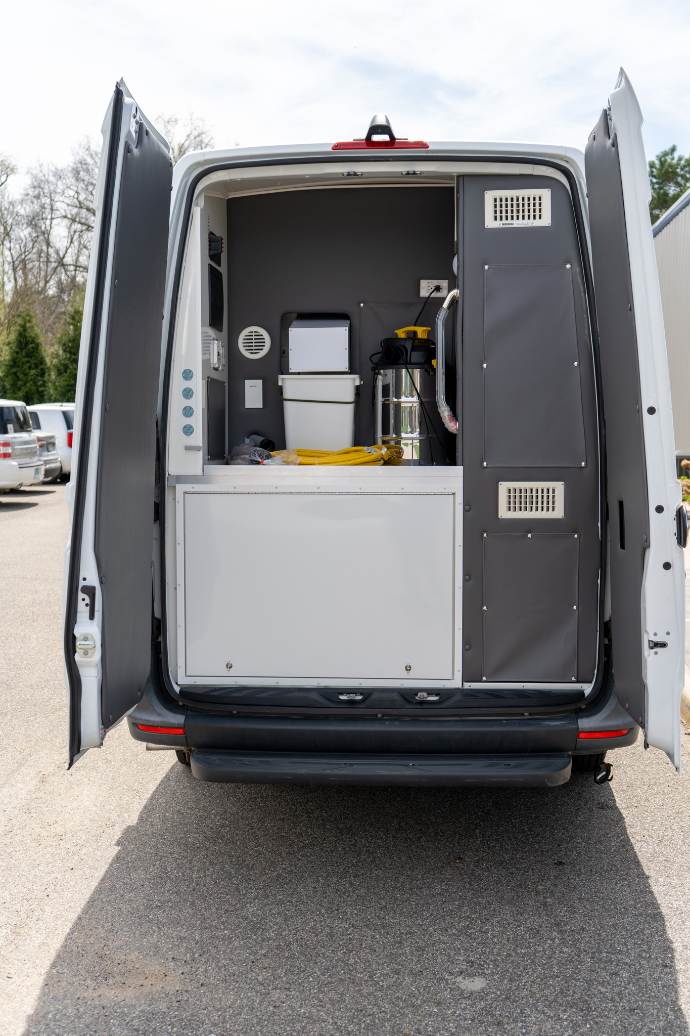 Open rear doors of a white work van showing a gray-fitout interior with cabinets, a white bin, yellow cables, and tools ready for service call