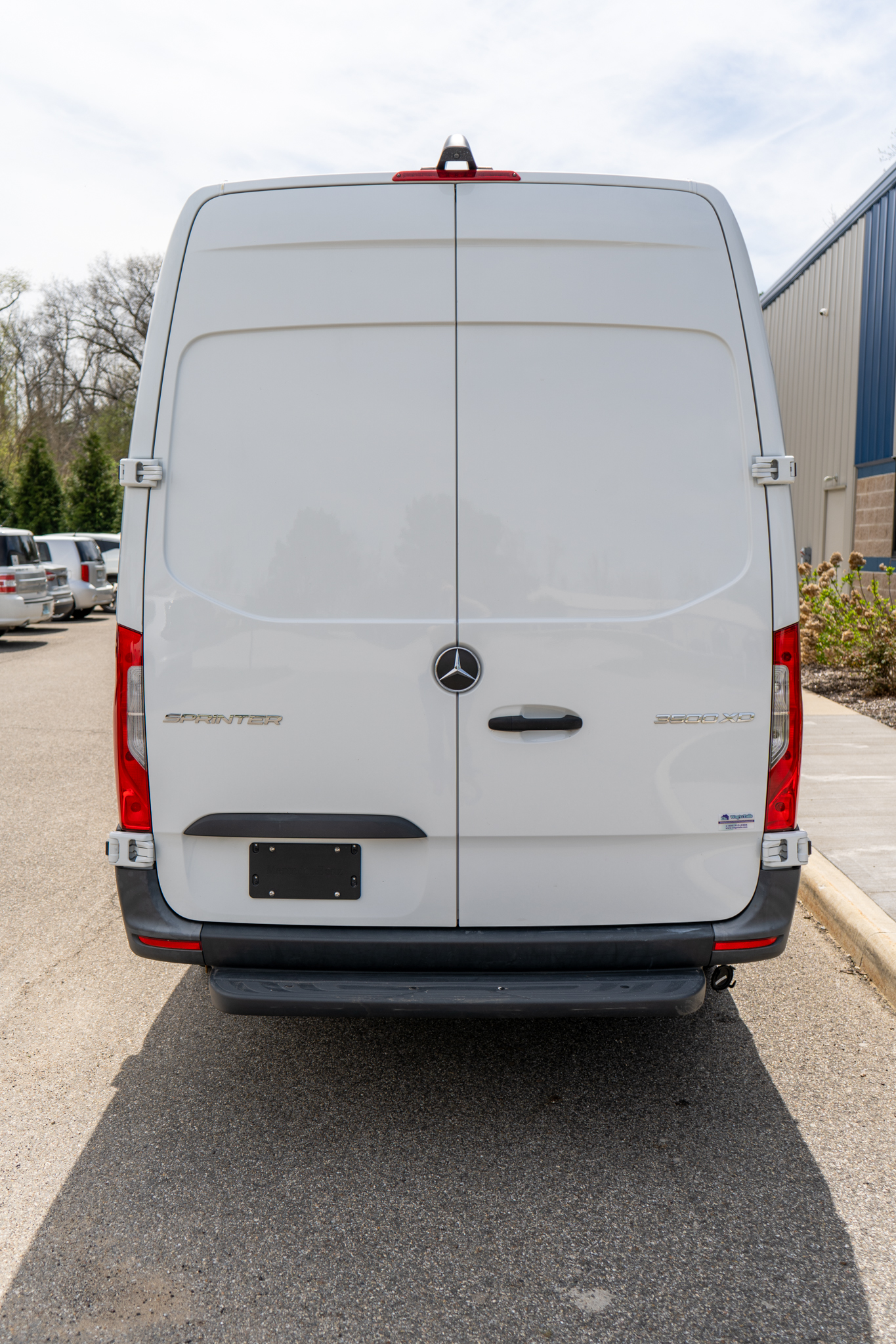 Rear view of a white Mercedes-Benz Sprinter van with double back doors and visible taillights on both sides in a parking lot.