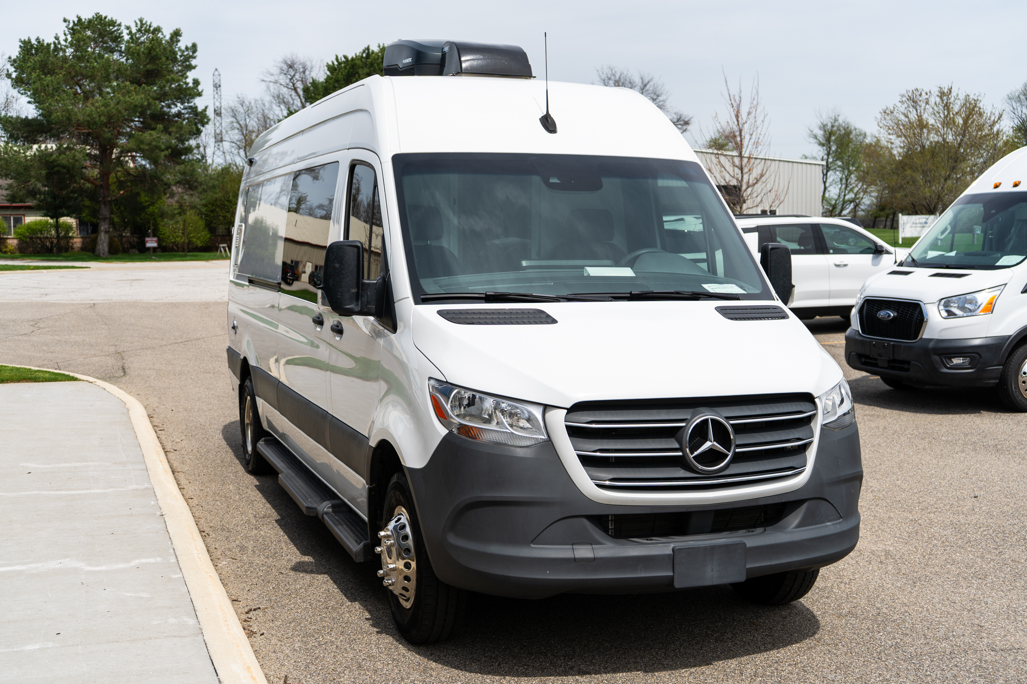 Front three-quarter view of a white Mercedes Sprinter-style van parked in a lot, with a rooftop cargo box visible on top.