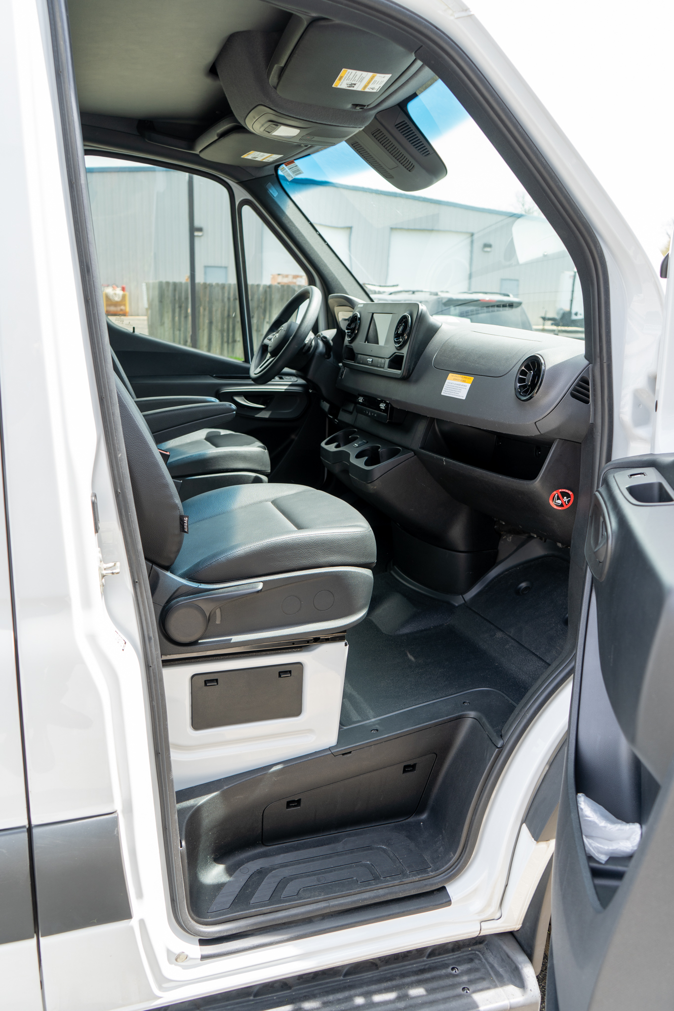 Driver’s cockpit of a white cargo van: front seat, steering wheel, center console, and dashboard with vents, door open.