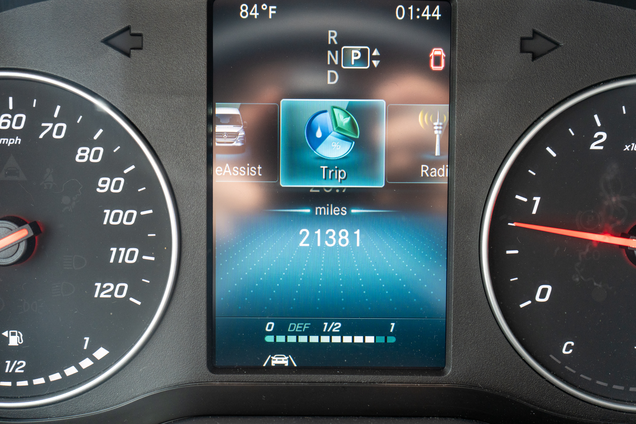 Center touchscreen shows Trip selected with nearby icons; time 01:44 and outside temp 84°F shown, gear in Park, odometer 21381 miles, surrounded by analog speedometer (left) and tachometer (right).