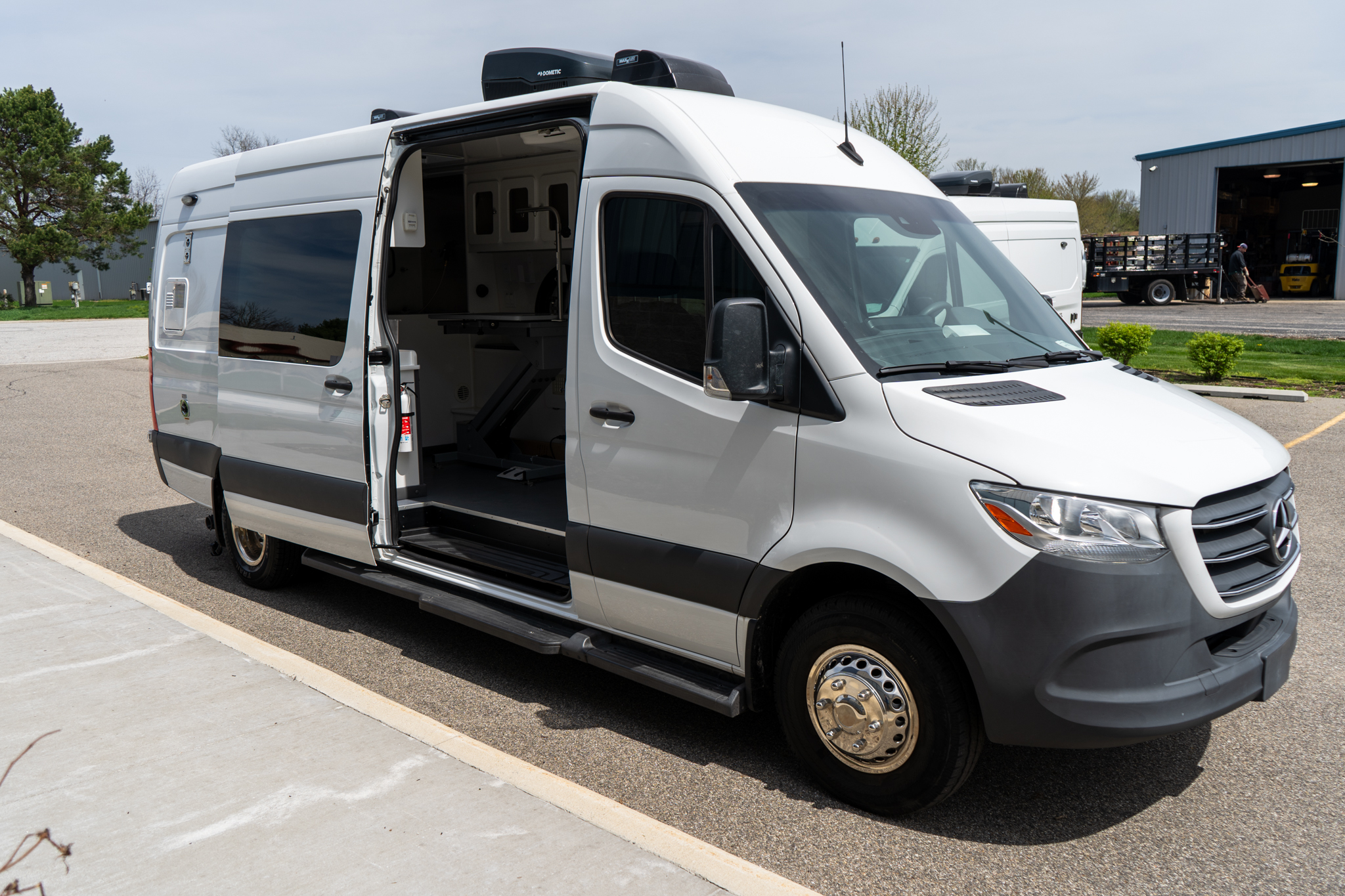 White cargo van with its side door open, revealing shelving and equipment inside, parked in a lot near a warehouse.