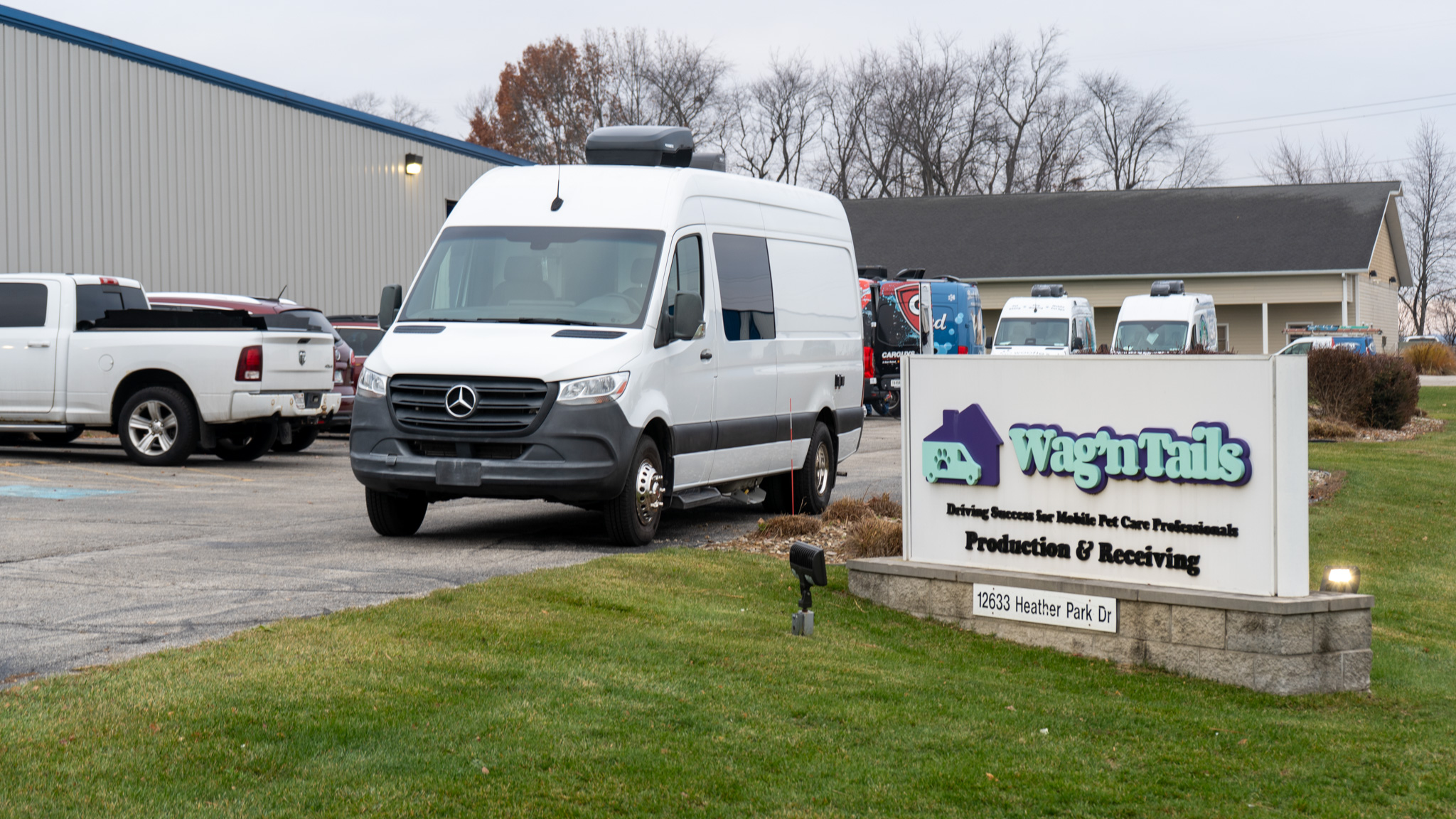 Front view of a white Mercedes van parked in a lot beside a Wag'n Tails sign, with other vehicles and a building in the background.