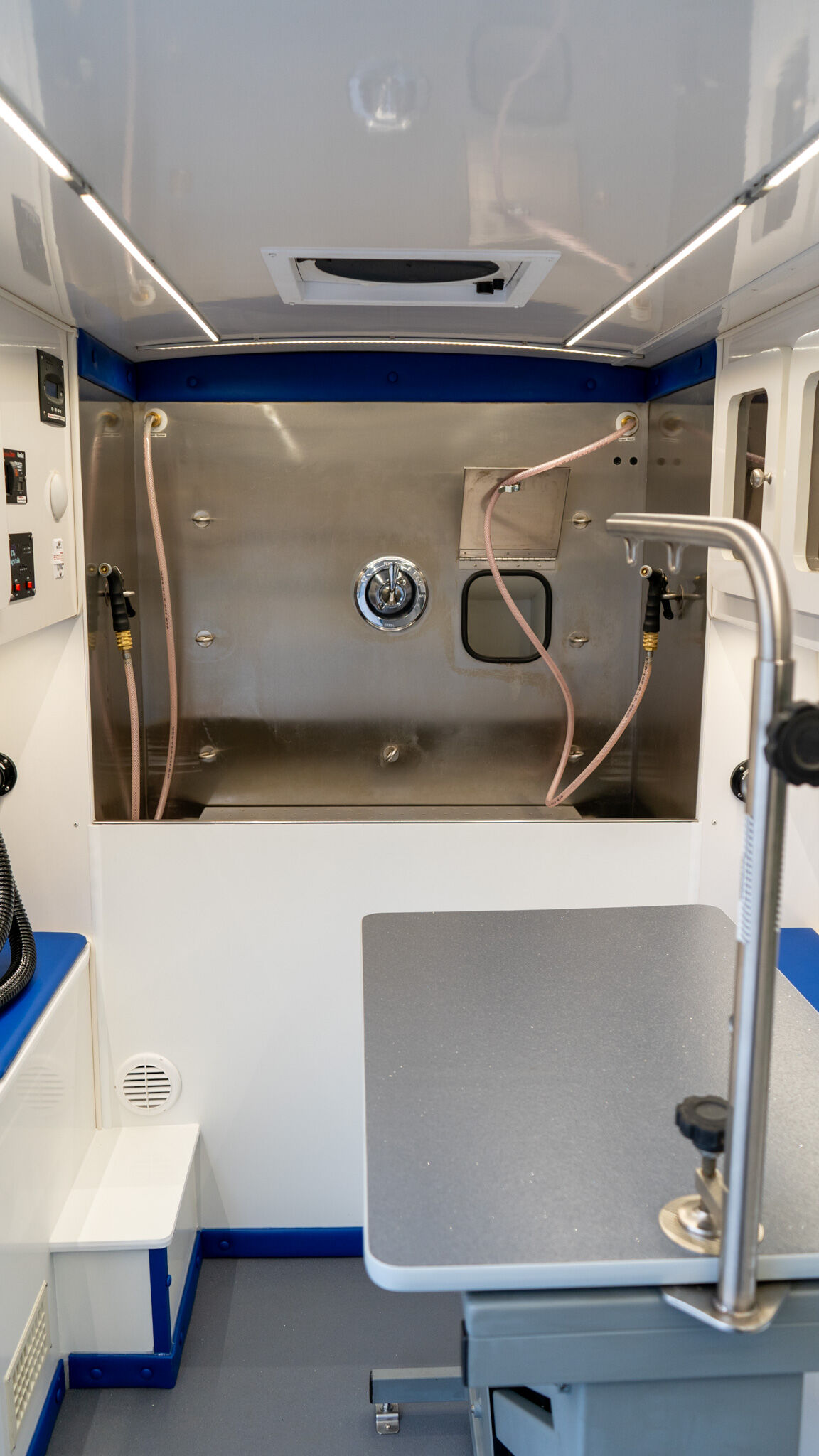Interior of a mobile medical clinic/ambulance with a stainless steel rear wall, pink hoses, control panel on the left, and a gray examination table nearby