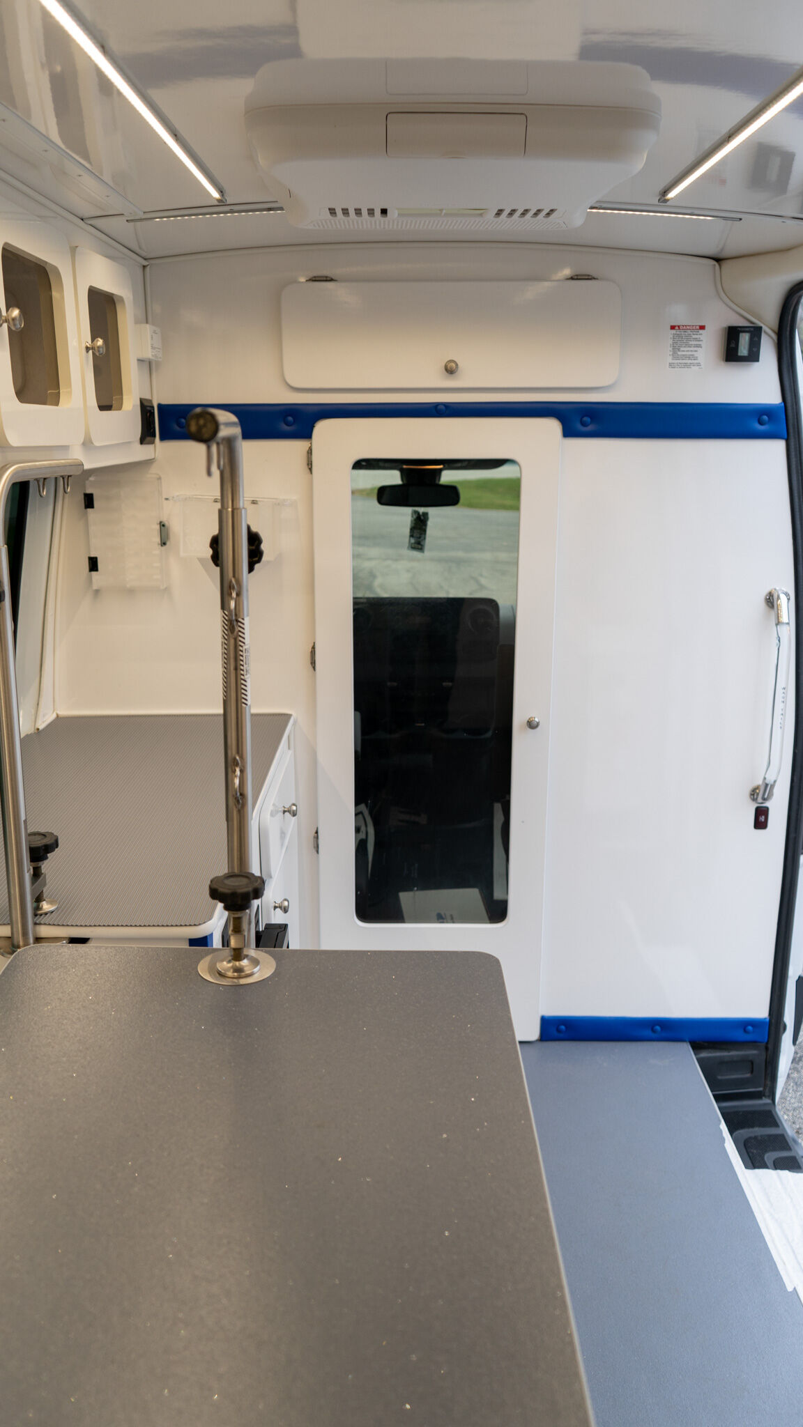 Interior view of a white ambulance cabin with a blue horizontal stripe, cabinets on the left, and a doorway at the back. A dark countertop is in the foreground.