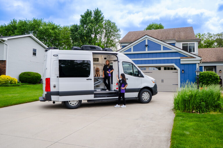 A white van with its side door open in a suburban driveway; a woman stands inside near a dog while a girl stands outside, with a blue house in the background.