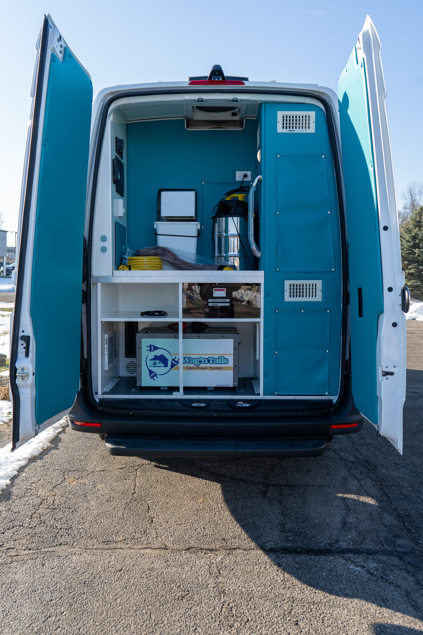 Open rear doors of a teal mobile service van, showing a stocked interior with shelves, electrical equipment, hoses, and a Wag'nTails CleanPower System unit.