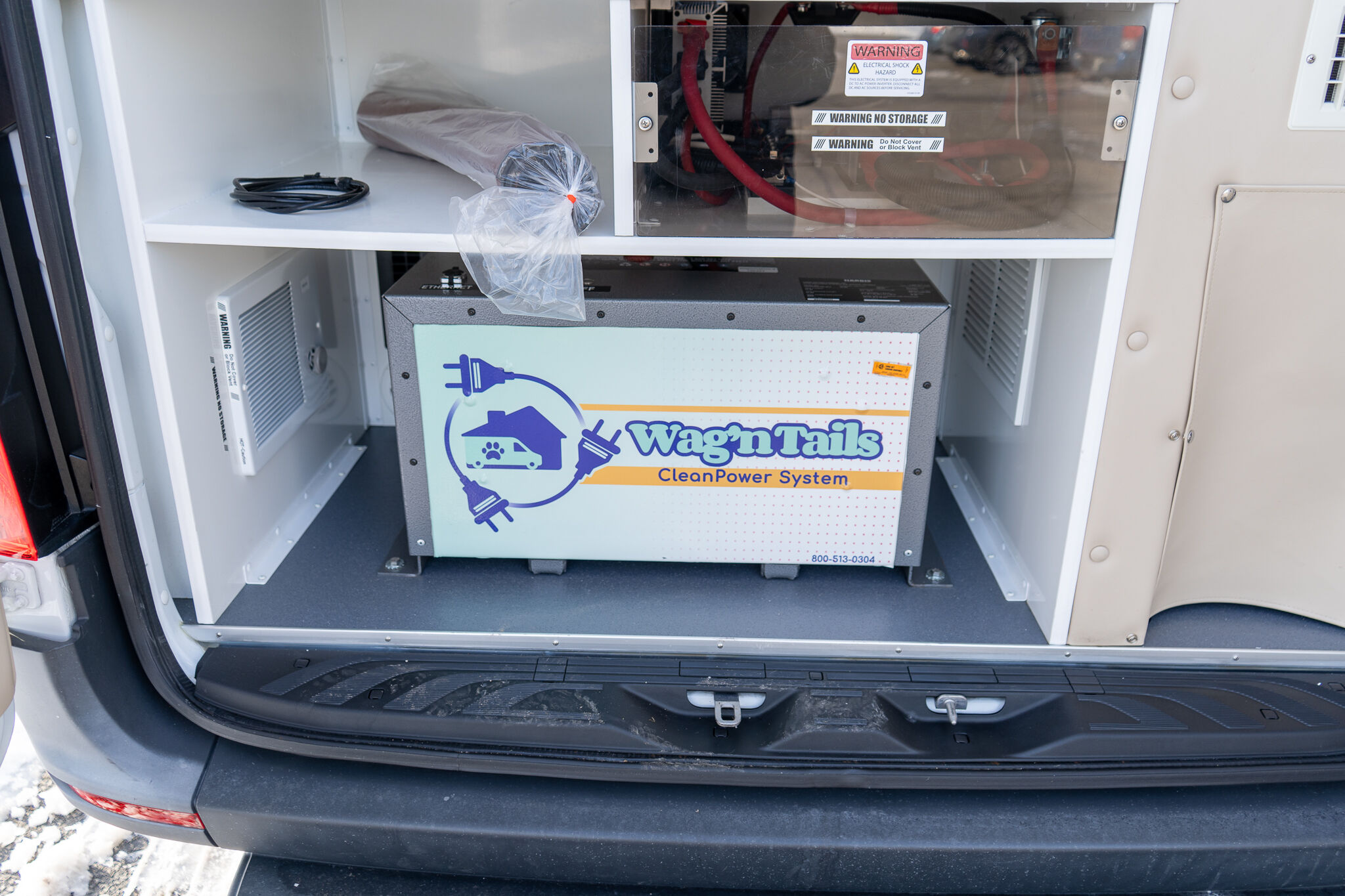 Open van cargo area showing a Wag'n Tails Clean Power System box with coiled cables and a plastic bag on top shelf.