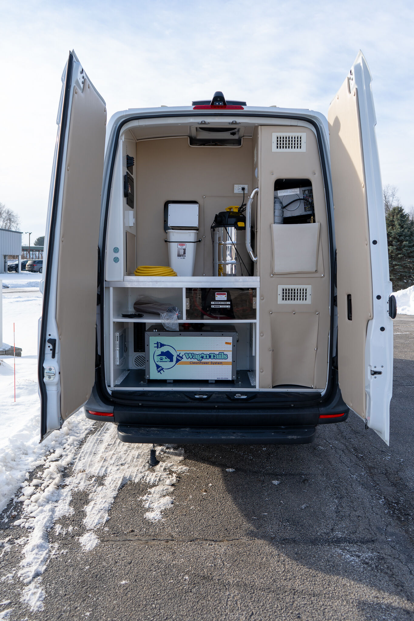 Open rear doors of a white service van revealing electrical equipment and storage shelves inside on a snowy lot.