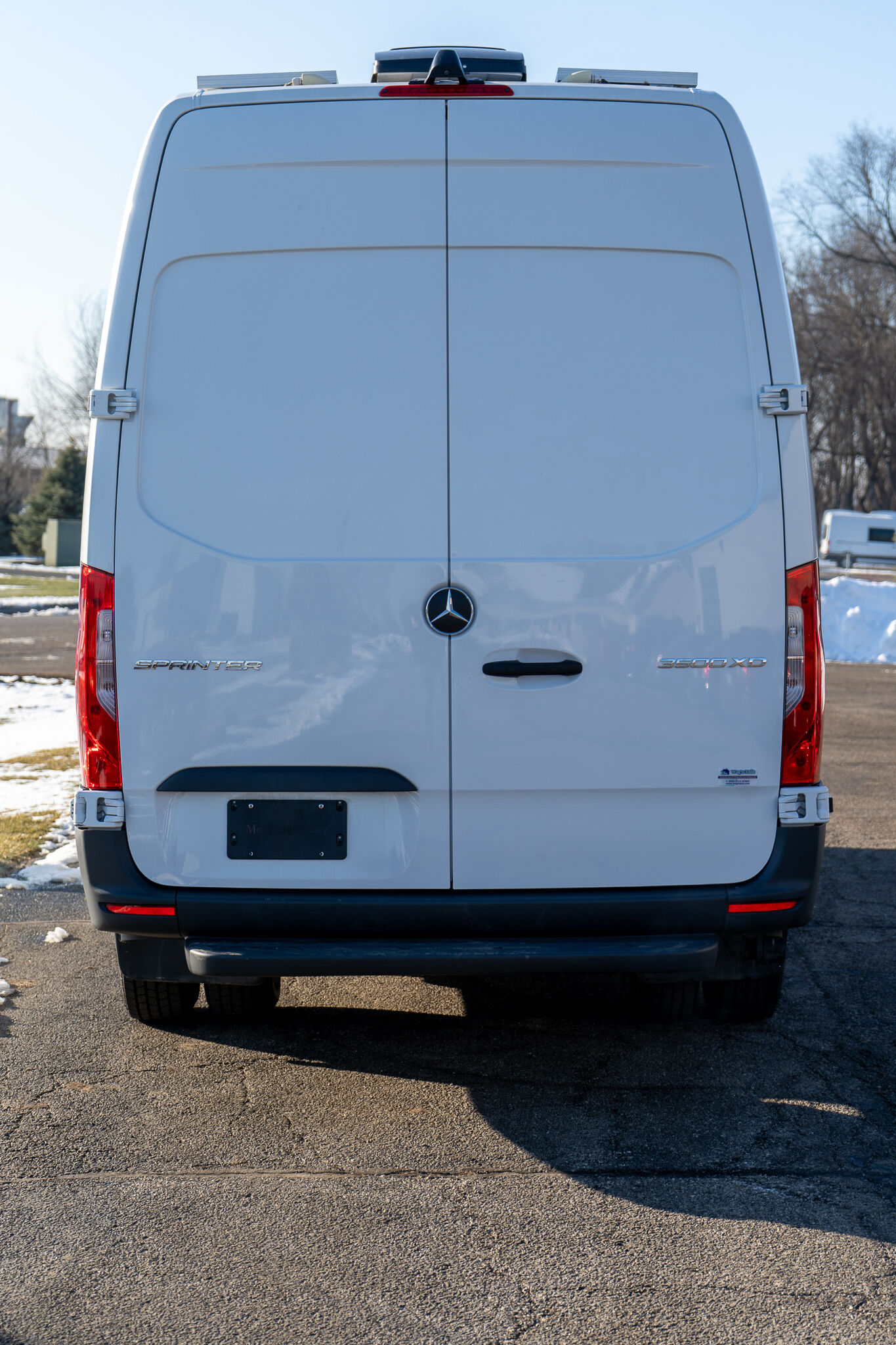 Rear view of a white Mercedes-Benz Sprinter van with two closed back doors, Mercedes badge centered, and model badges SPRINTER and 3500 XD on the right; red taillights visible in a parking lot with patches of snow on the ground.
