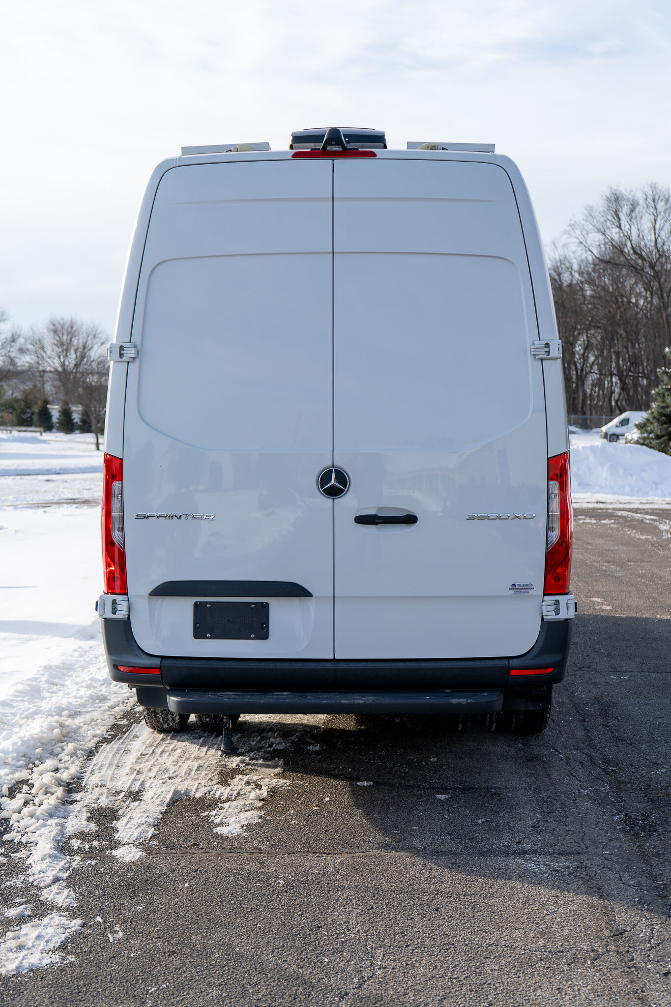 Rear view of a white Mercedes-Benz Sprinter van parked on a snow-covered road,/: visible Mercedes emblem and model badge on the back doors