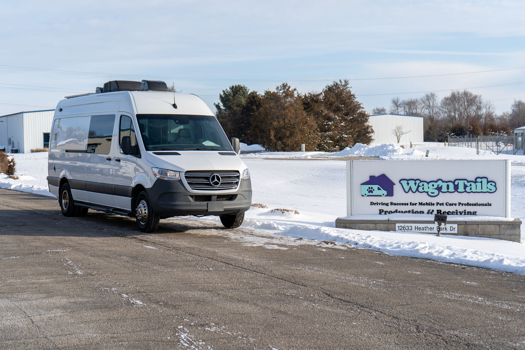 White Mercedes Sprinter van parked on a snowy lot beside a Wag'n Tails sign and industrial buildings in the background.