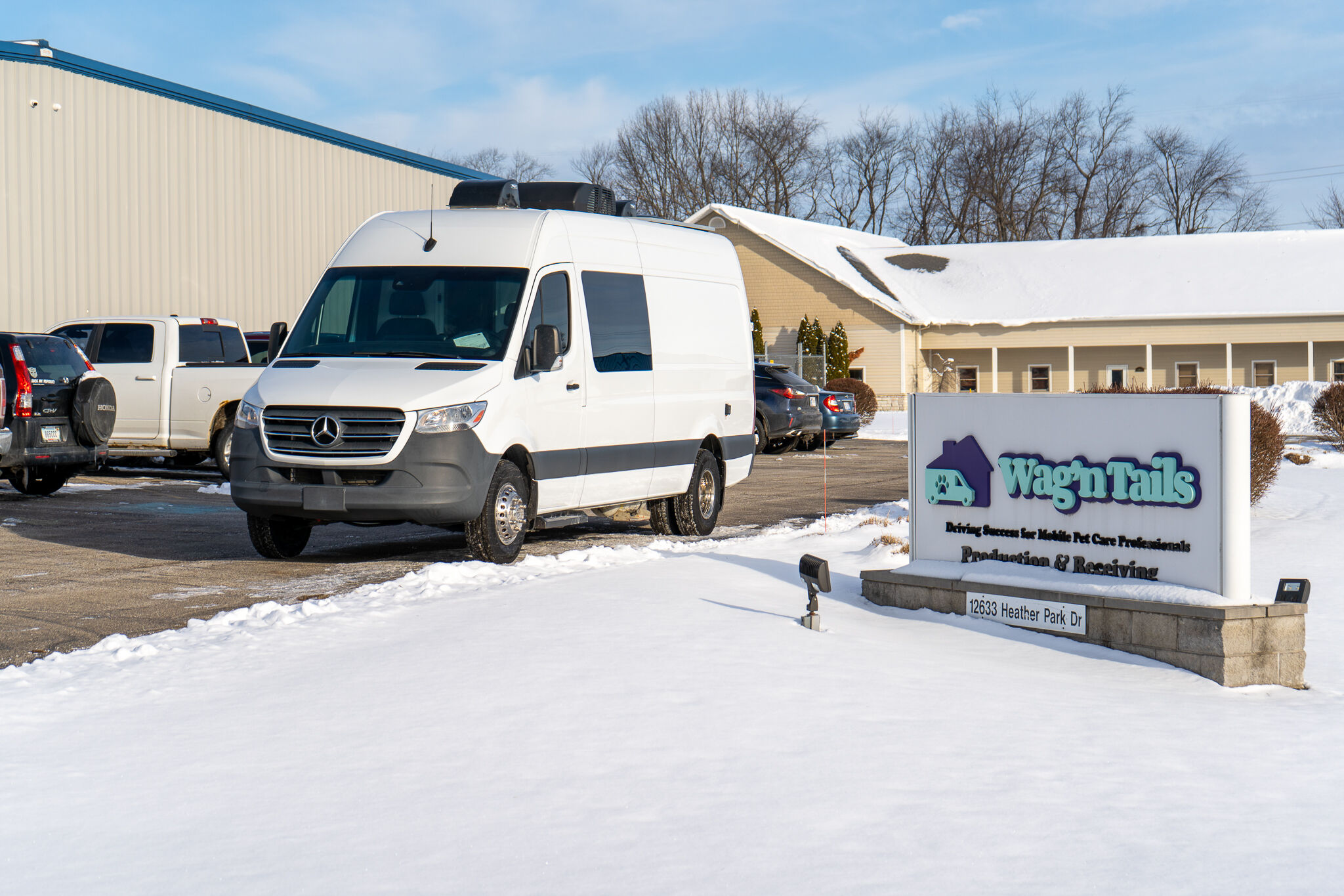White Mercedes Sprinter-style van parked in a snow-covered parking lot next to a Wag'n Tails sign outside a beige commercial building.