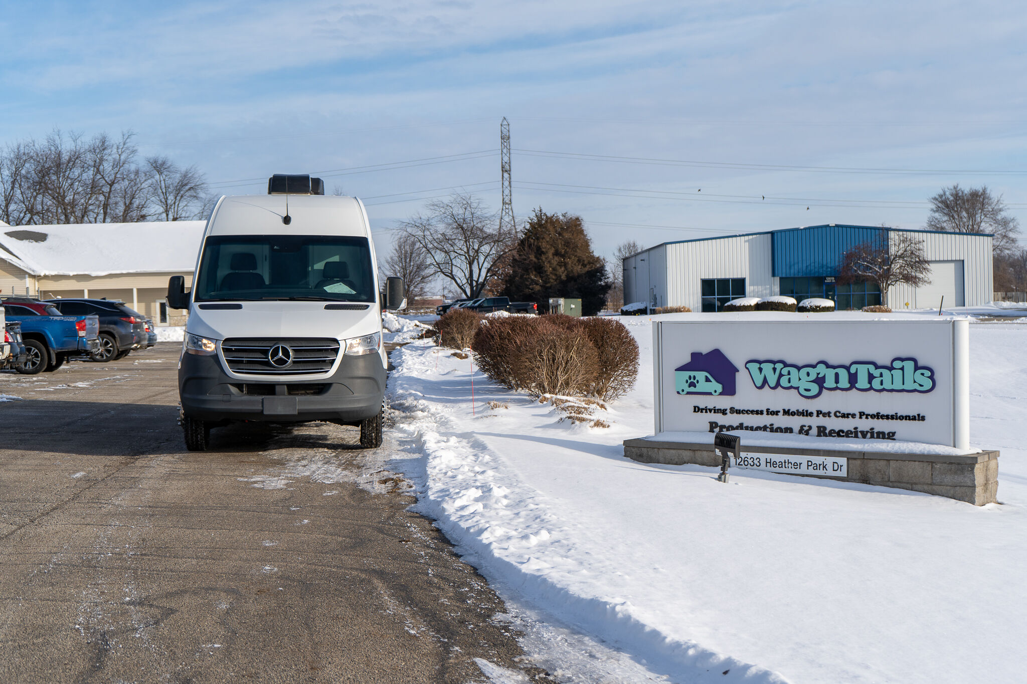 White Mercedes van in a snowy parking lot near a business sign that reads 'Wag n Tails' and a blue-and-gray industrial building in the background.