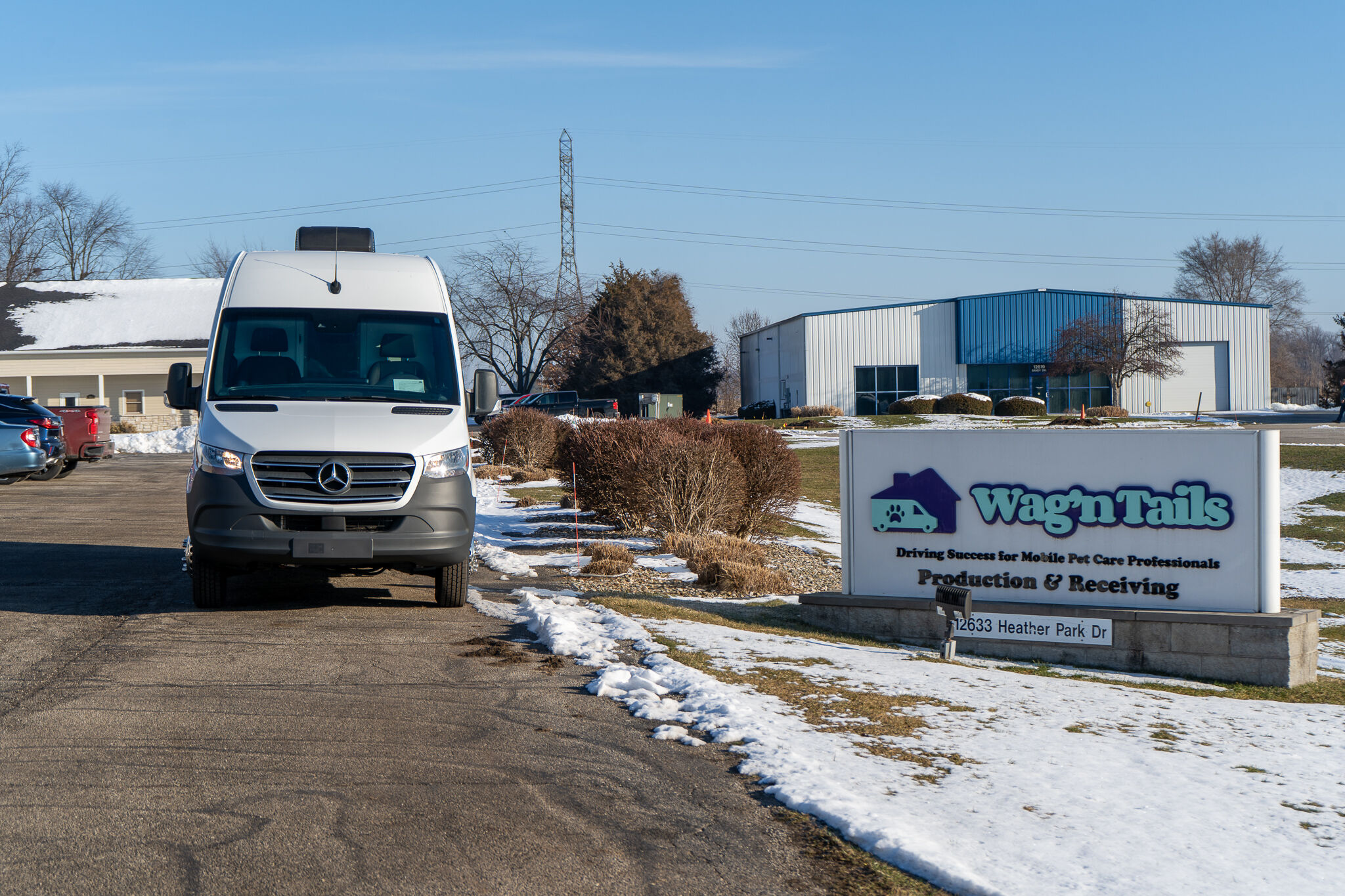 White Mercedes van parked in a lot in front of a Wag'n Tails pet care facility on a clear winter day, with snow along the edges of the pavement and a large sign visible nearby.
