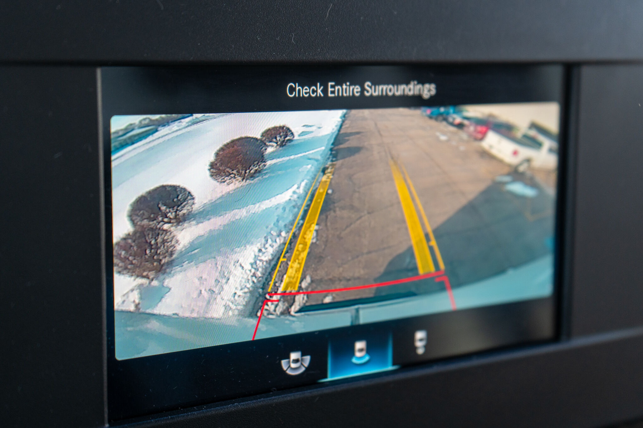 Rear-view camera display with yellow/red guide lines in a snowy parking lot, showing nearby trees and a vehicle in view.