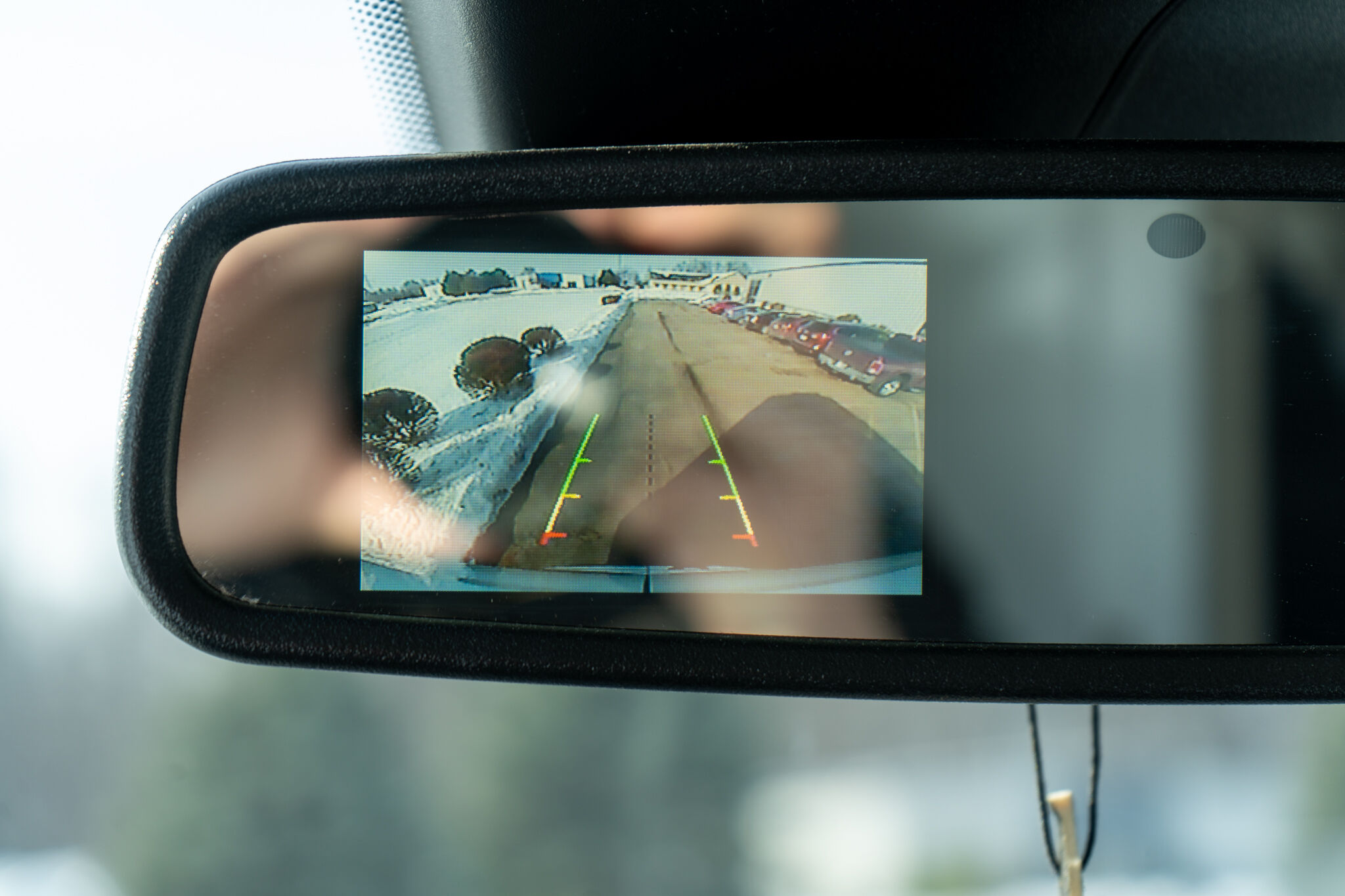 Rearview mirror displaying a car's backup camera feed of a parking lot with guiding colored lines on the screen.