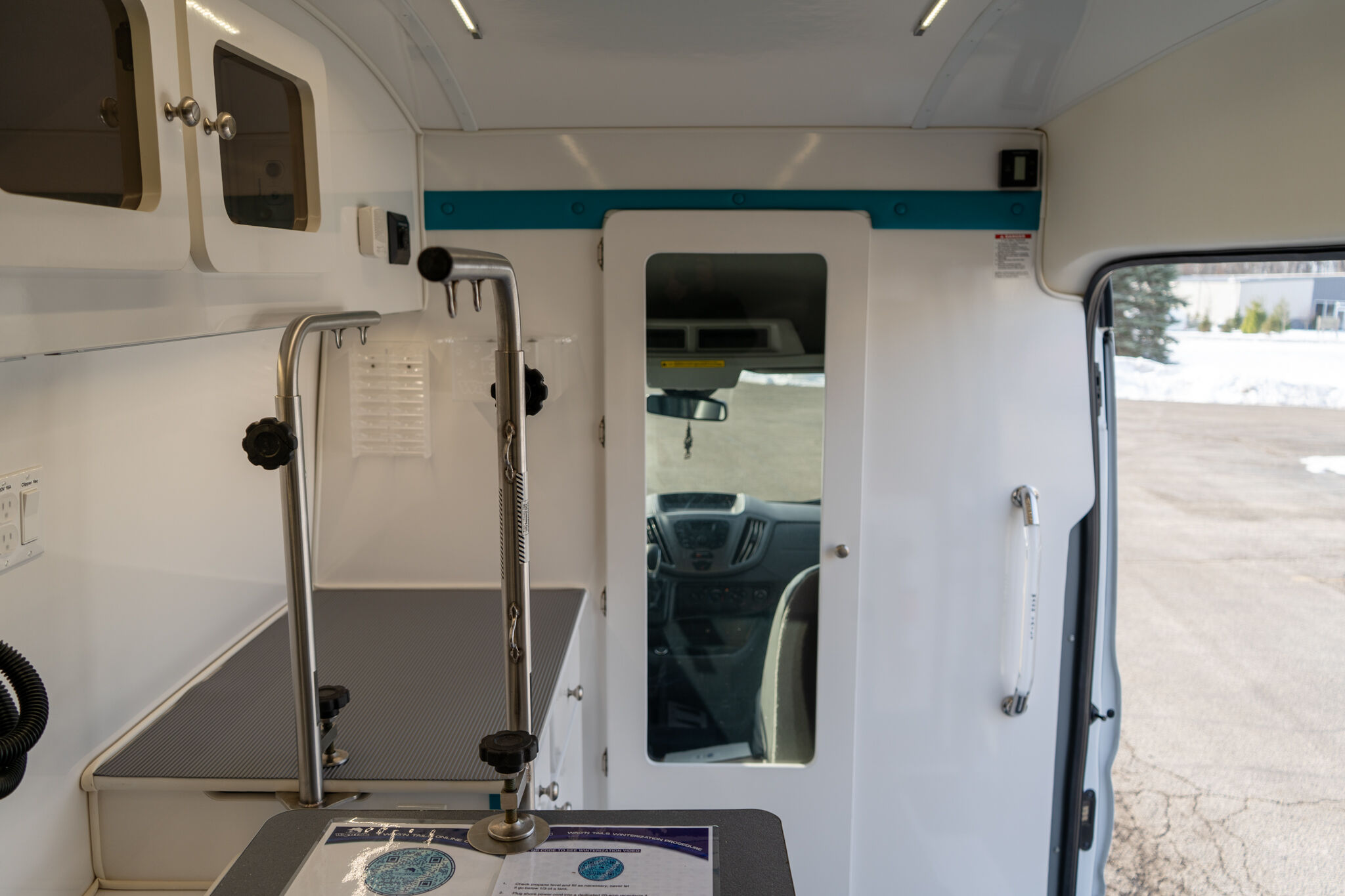 Interior of a white service van with metal rails, a working counter, and an open doorway to the cab, light fixtures above, and a snowy outdoor scene visible through the side door